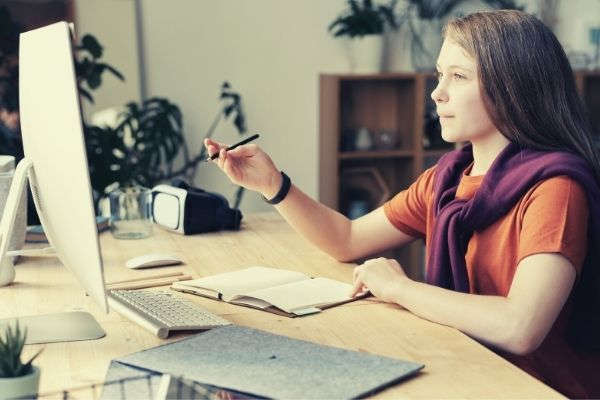 girl-holding-pencil-while-looking-at-imac-netiquette-rules-wearing-dark-orange-shirt-and-sweater-wrapped-around-her-shoulder
